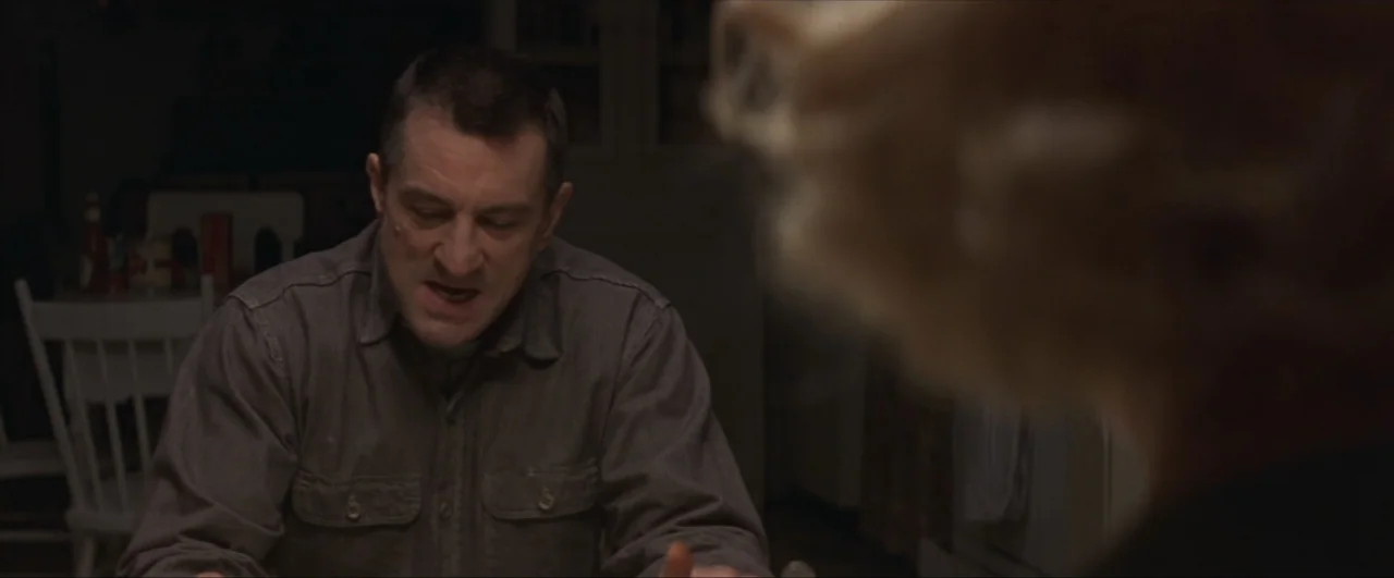 This Boy's Life (1993) movie still: medium — A dimly lit room contains a white wooden chair and a dining table. A man in a dark grey shirt sits at…
