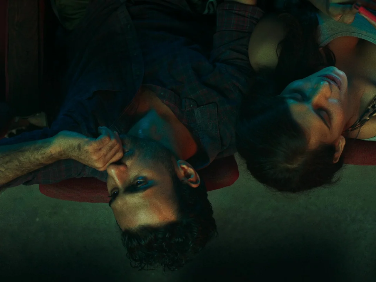 Joyland (2022) movie still: medium — A dark, cramped interior space illuminated by the cold blue light of a screen. A man and a woman lie together…