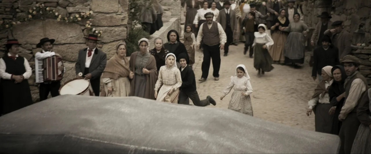 Fatima (2020) movie still: wide — A bustling 1900s village street is lined with weathered stone walls and historic architecture. A diverse crowd of…