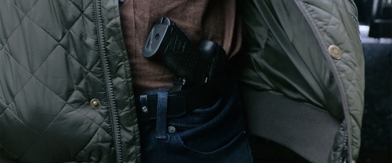 The Gentlemen (2020) movie still: closeup — A dark green quilted jacket is open to reveal a handgun tucked into a belt. The weapon rests against a…