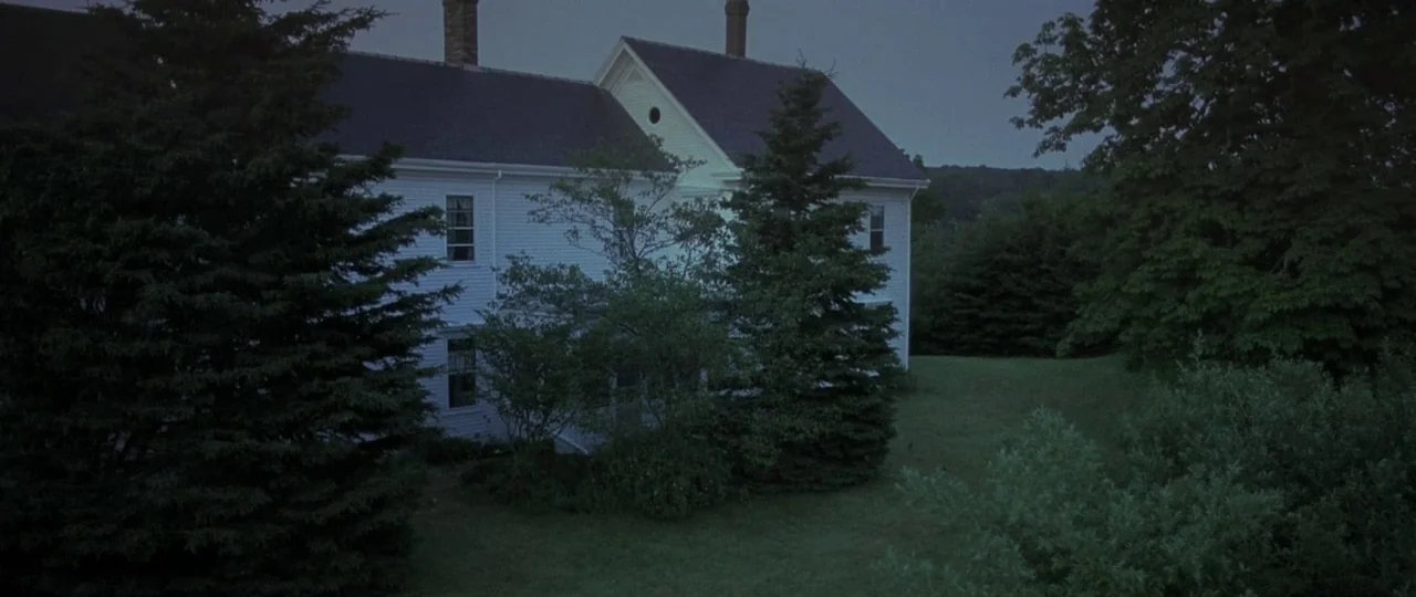 In the Bedroom (2001) movie still: establishing — A white clapboard farmhouse is surrounded by dense evergreen trees and a grassy yard. The house…