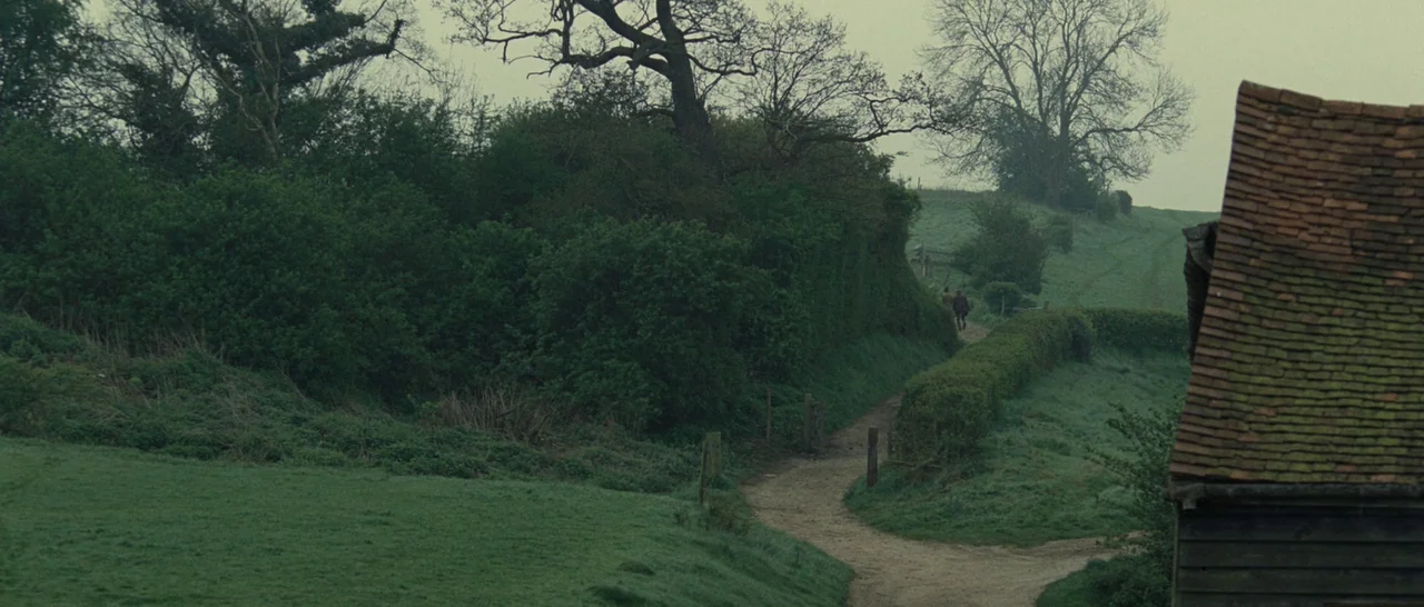 Never Let Me Go (2010) movie still: wide — A rural dirt path winds through lush green hills and dense hedgerows. A small rustic wooden outbuilding…
