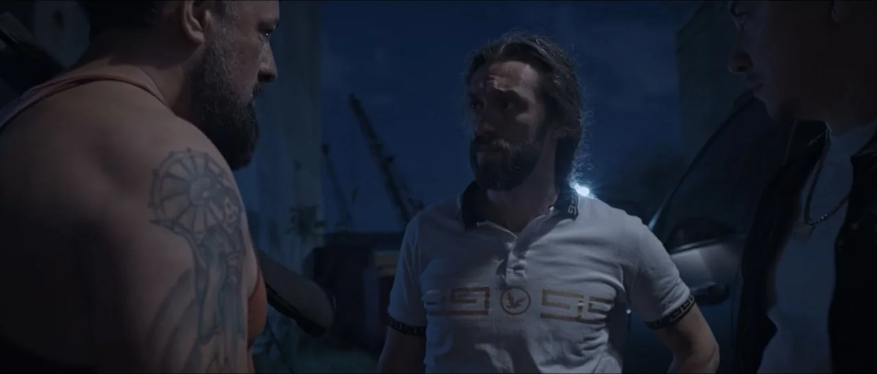 Gunman (2025) movie still: medium — Three men stand outdoors in an industrial setting at night near parked vehicles. They appear to be in a quiet…
