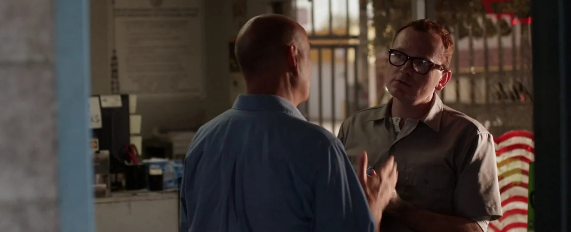 Cheap Thrills (2013) movie still: medium — A man in a tan shirt speaks to a bald man in a dusty room. The interior appears to be a cluttered…