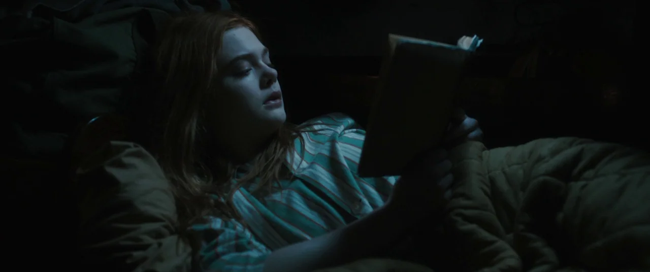Ginger & Rosa (2012) movie still: closeup — A person lies in a dark room under a warm blanket at night. A young girl with long red hair reads a book…