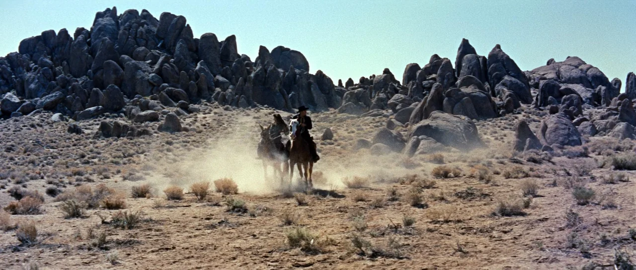 Comanche Station (1960) movie still: wide — A vast, arid desert landscape features prominent grey rock formations and sparse, dry brush. Two riders…