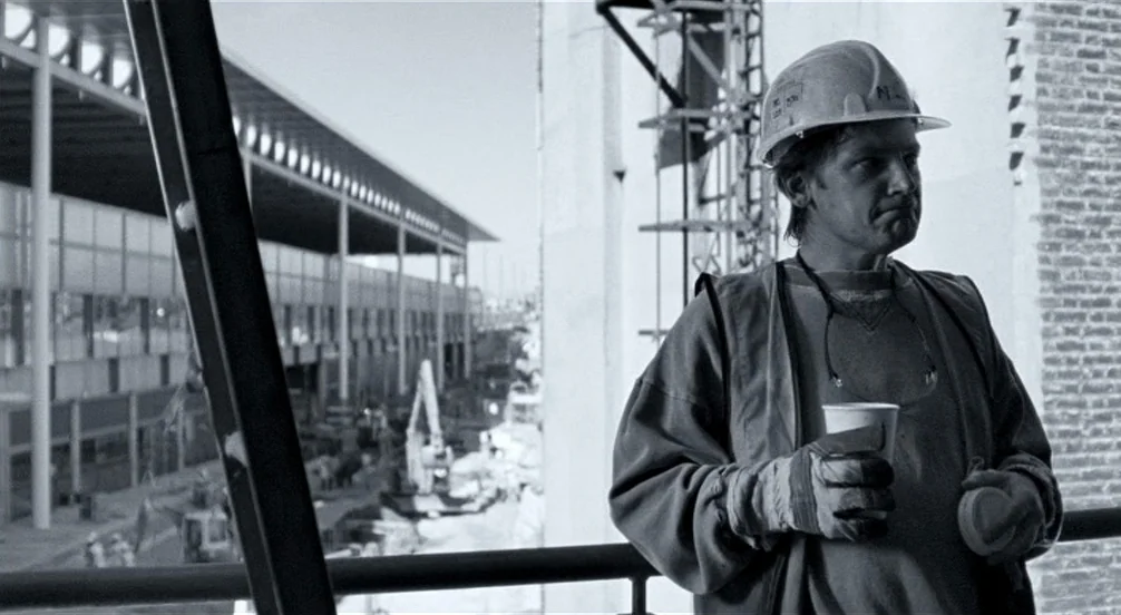 Somers Town (2008) movie still: medium — A construction worker stands on a scaffold overlooking a large building project. He is holding a paper cup…