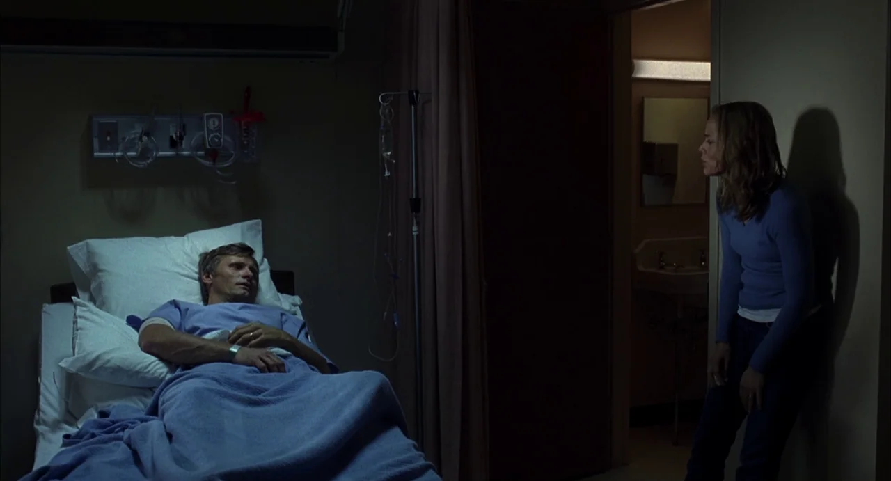 A History of Violence (2005) movie still: wide — A dimly lit hospital room features a man resting in bed. A woman stands in the shadows of the…