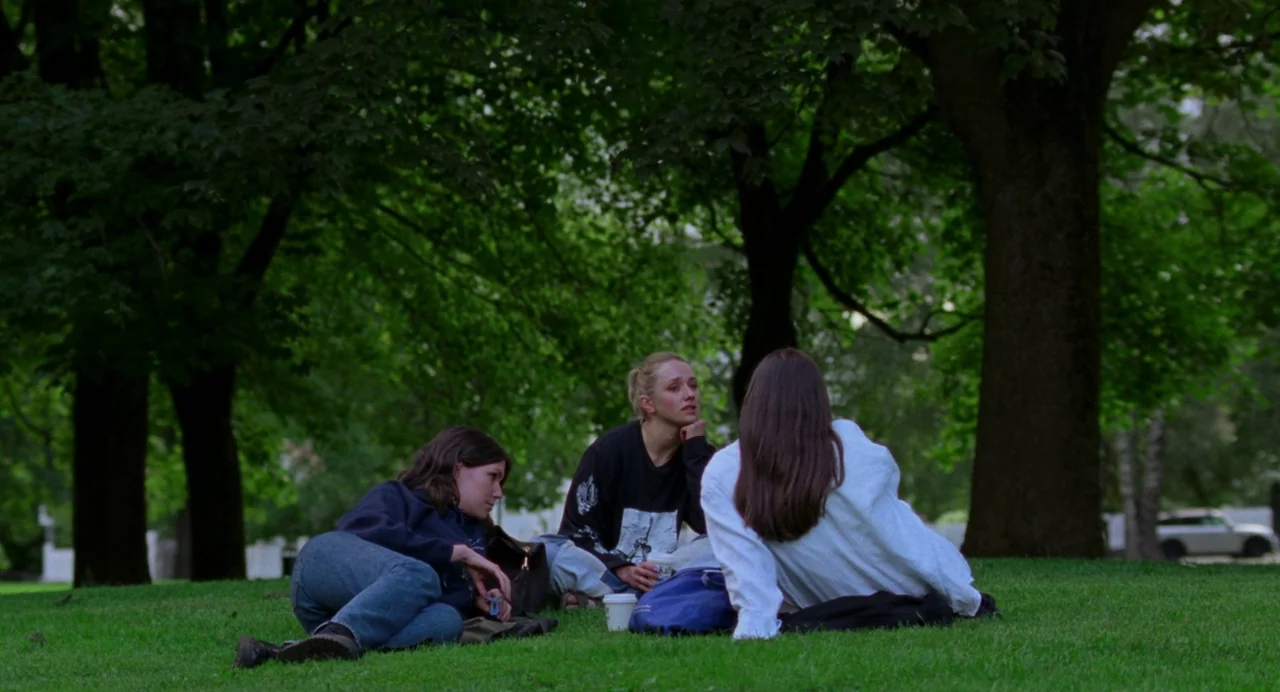 Sick of Myself (2022) movie still: fullbody — Three young women sit together on a manicured lawn beneath large, shaded trees. They are positioned in…