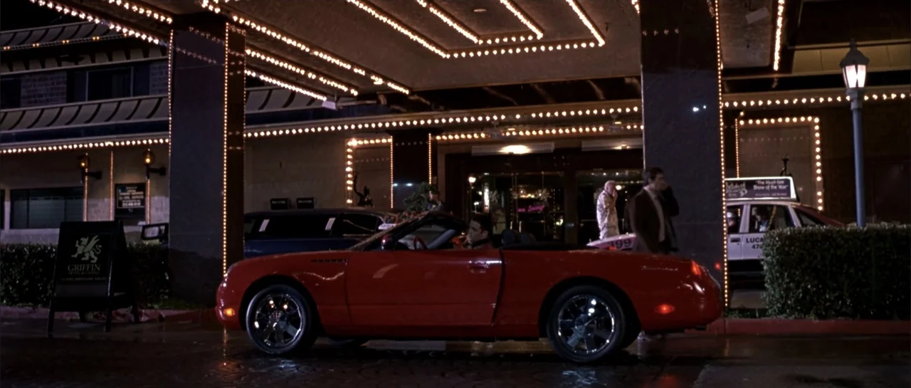 Shade (2003) movie still: wide — A night exterior shot of a hotel entrance illuminated by numerous warm lightbulbs. A man sits in a bright red…