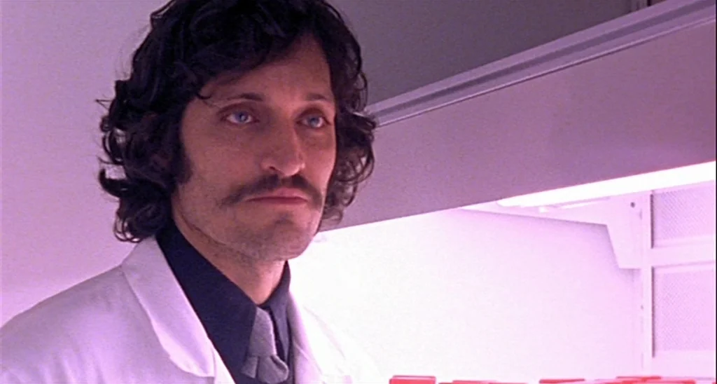 Trouble Every Day (2001) movie still: closeup — A man in a lab coat stands in a brightly lit, sterile room. He looks forward with a neutral…
