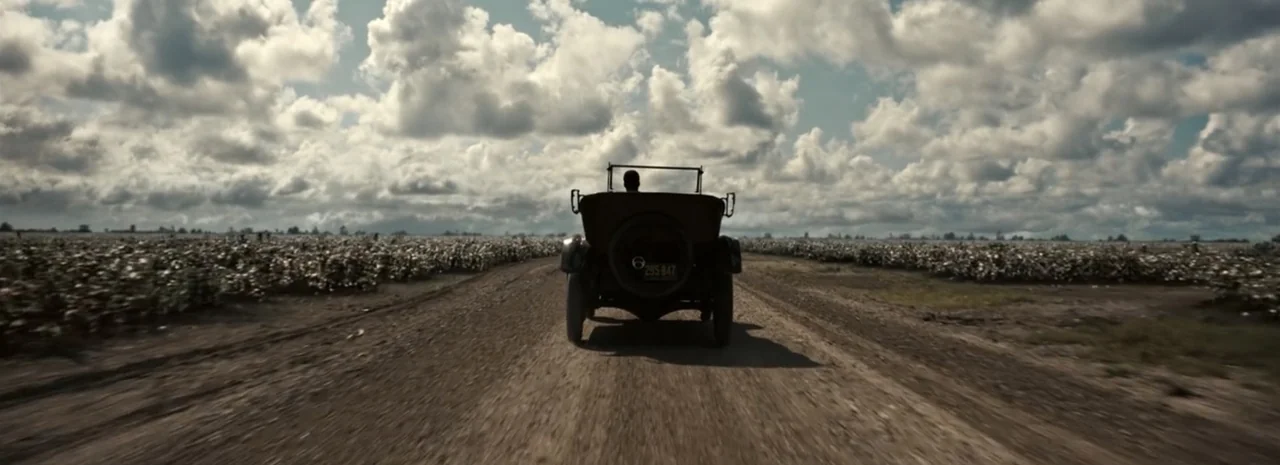 Sinners (2025) movie still: wide — A vast, open rural landscape with endless fields stretches toward a cloudy horizon line. A vintage 1930s…