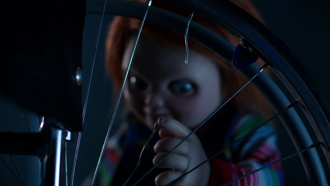 Cult of Chucky (2017) movie still: closeup — A doll sits in a dark space, partially obscured by a bicycle wheel. The doll's hand extends toward a…