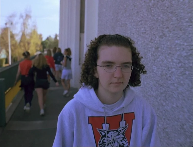 Elephant (2003) movie still: medium — An outdoor concrete walkway outside a building with students walking in the background. A teenage girl in a…