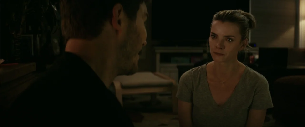 The Tomorrow War (2021) movie still: medium — A dark, dimly lit domestic space features a couch and home entertainment equipment. A woman sits…