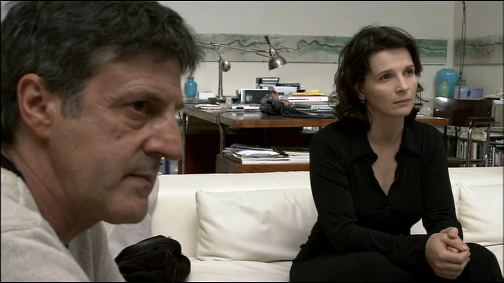 Caché (Hidden) (2005) movie still: medium — A modern living room features a white sofa and a cluttered desk behind it. A man and a woman are seated…