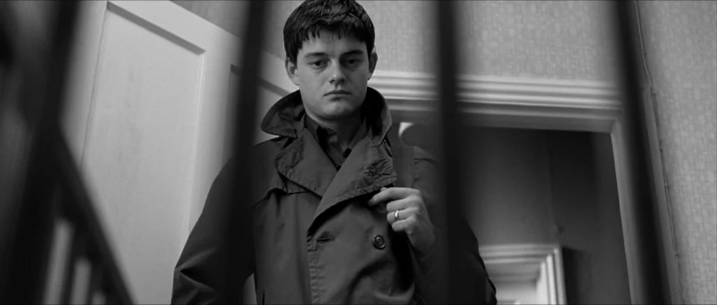 Control movie still: medium — A residential staircase features white paneling and a vertical black railing in the foreground. A young man wearing a…