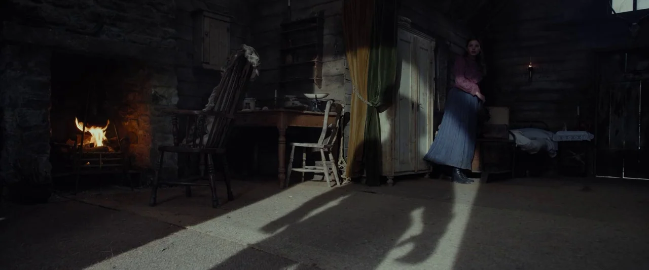 Apostle (2018) movie still: wide — A dimly lit log cabin features dark wood walls and a stone fireplace. A woman stands in a bright light shaft cast…