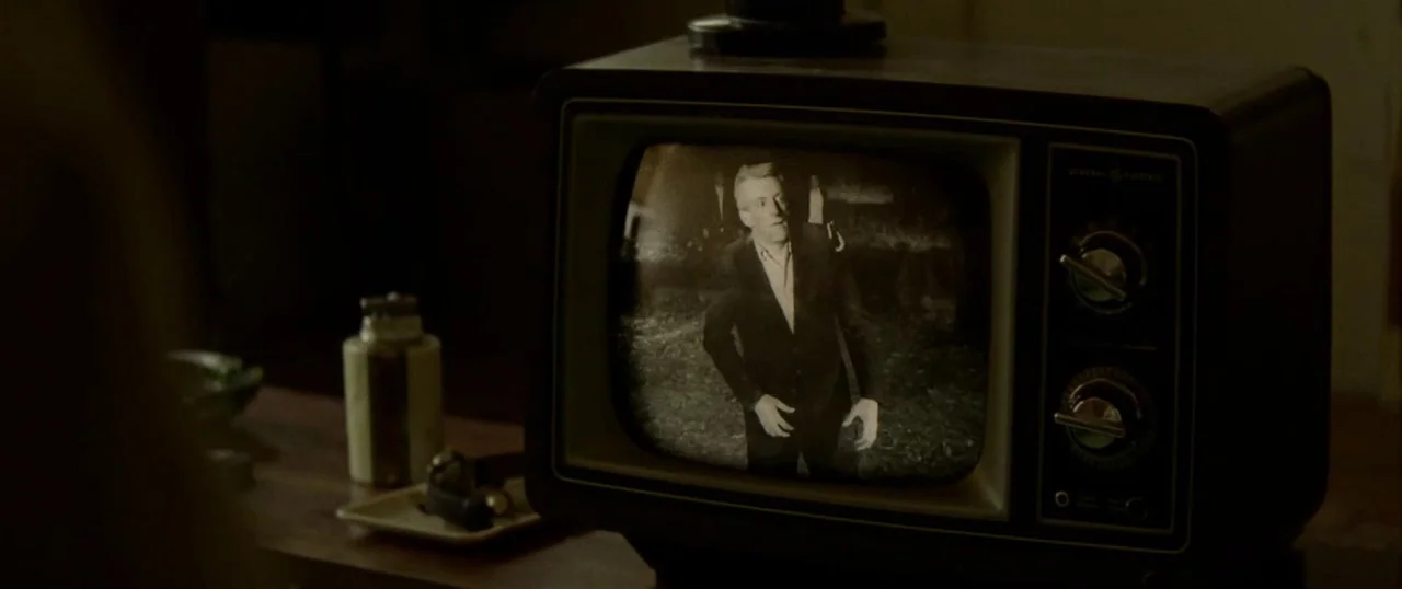 Low Down (2014) movie still: medium — A vintage boxy television sits on a wooden surface in a dark room. The screen displays a black-and-white image…