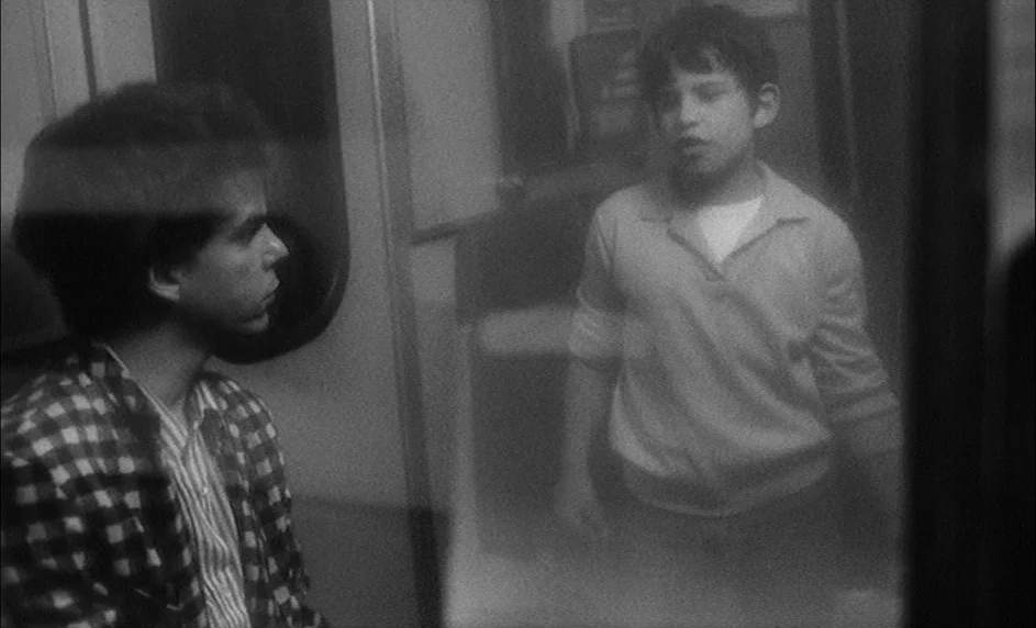 Boy Meets Girl (1984) movie still: medium — A dimly lit interior of a train carriage with metal panels and reflections visible. Two young males are…