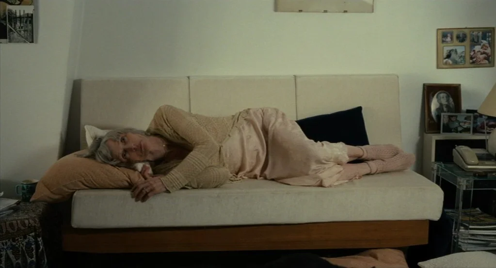 Things to Come (2016) movie still: wide — A living room interior features a white daybed against a plain wall with framed photos. An elderly woman…