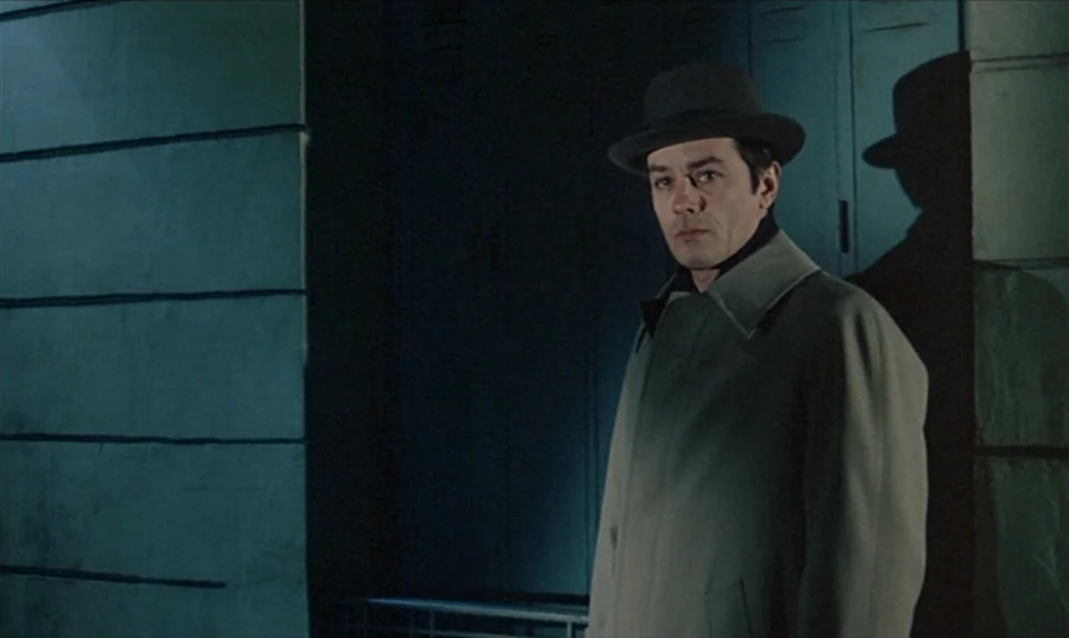 Mr. Klein (1976) movie still: medium — A man stands on a sidewalk next to a dark blue industrial or apartment wall. He is illuminated by a single…