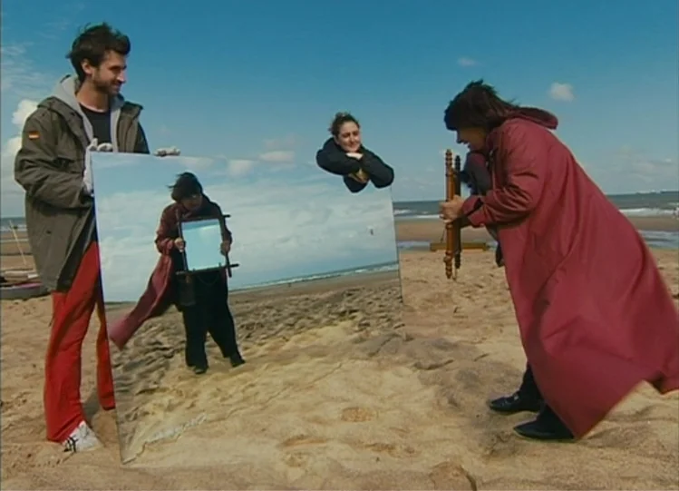 The Beaches of Agnès (2008) movie still: fullbody — A sunny beach with light sand and a bright blue sky. Three people manipulate a large rectangular…