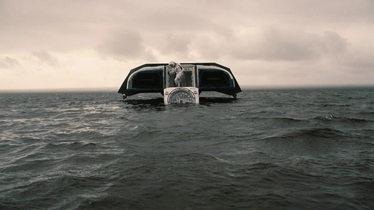 Interstellar (2014) movie still: wide — A vast, calm ocean stretches toward a horizon beneath a thick, overcast grey sky. A futuristic lander sits…