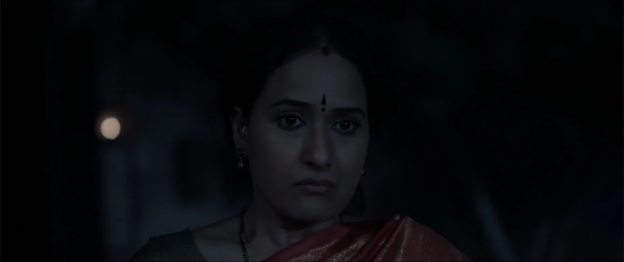 Cheekatilo (2026) movie still: closeup — A dark, dimly lit interior space with minimal architectural features visible in the shadows. A South Asian…