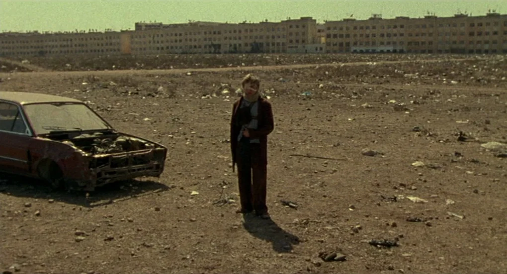 Furia (1999) movie still: wide — A vast, barren lot strewn with scattered debris and trash sits under clear skies. A young man stands beside the…