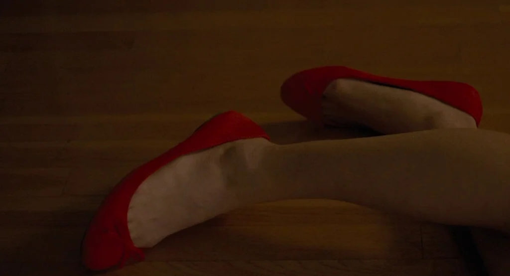 Ruby Sparks (2012) movie still: closeup — Dark wood floorboards stretch across the frame in dim, low-key lighting. A pair of red ballet flats rests…
