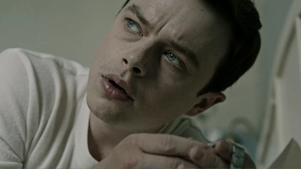 A Cure for Wellness (2017) movie still: closeup — A spare, dimly lit bedroom features pale, neutral walls and soft window lighting. A young man lies…