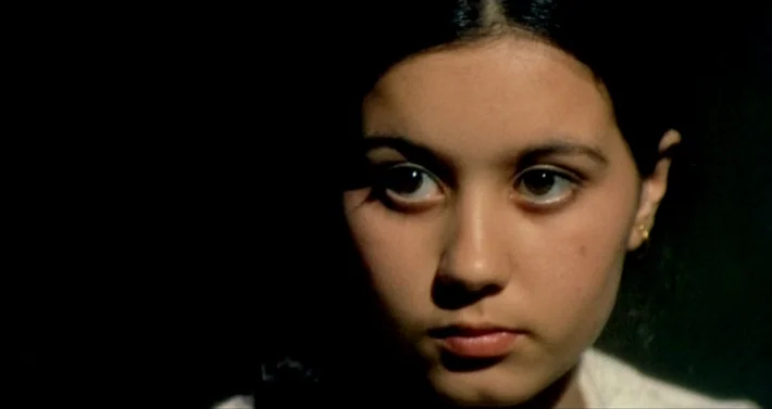 Blood For Dracula (1974) movie still: closeup — A teenage girl is framed against a solid black background. She gazes directly forward with an…