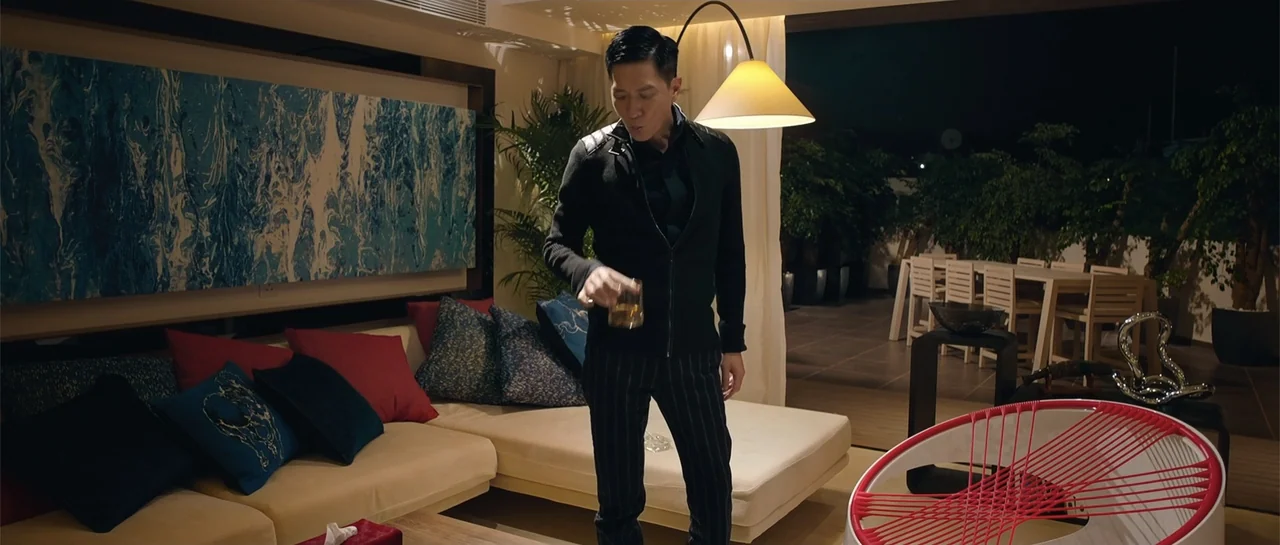 Line Walker (2016) movie still: medium — A modern living room contains a beige sofa with colorful cushions and a patio. An East Asian man in a black…