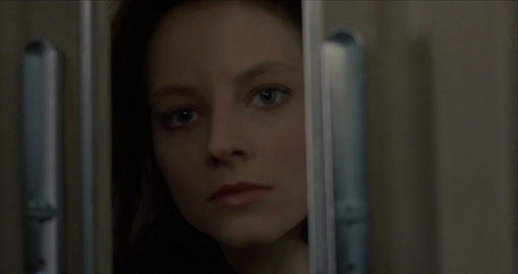 The Silence Of The Lambs (1991) movie still: closeup — A close-up view through a vertical gap between two metal industrial doors. A young woman with…
