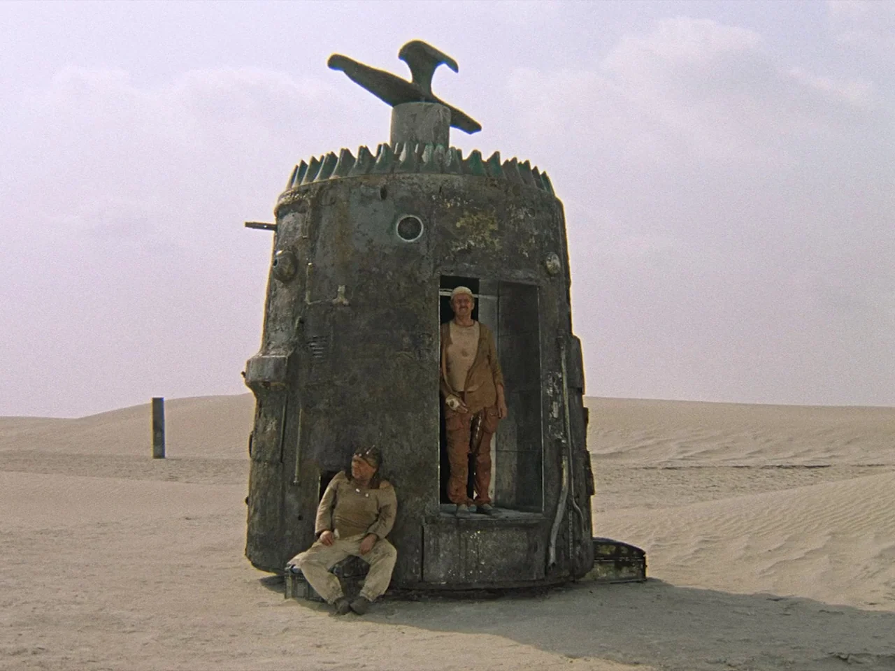 Kin-dza-dza! (1986) movie still: wide — A vast, barren desert contains a singular, oxidized metallic structure with a protruding top piece. Two men…