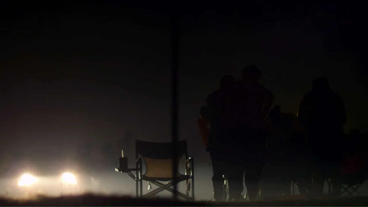 Rich Hill (2014) movie still: wide — A dark outdoor space is illuminated by two distant lights near the horizon. A group of people stands in…