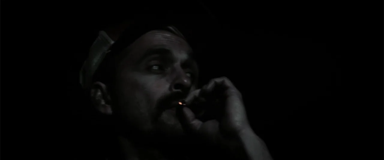 At Dawn (2013) movie still: closeup — A dark space provides a backdrop for a man illuminated by a cigarette ember. The subject sits in deep shadow…