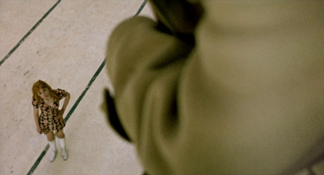 Deep End (1970) movie still: fullbody — A light-colored gymnasium floor features painted dark boundary lines. A woman stands looking upward from a…