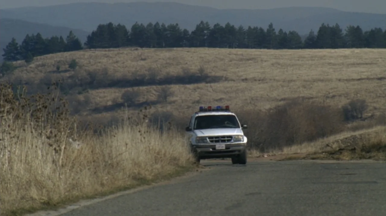 The Shepherd (2023) movie still: wide — A white police SUV travels on a paved road through a dry, hilly landscape. The vehicle drives toward the…