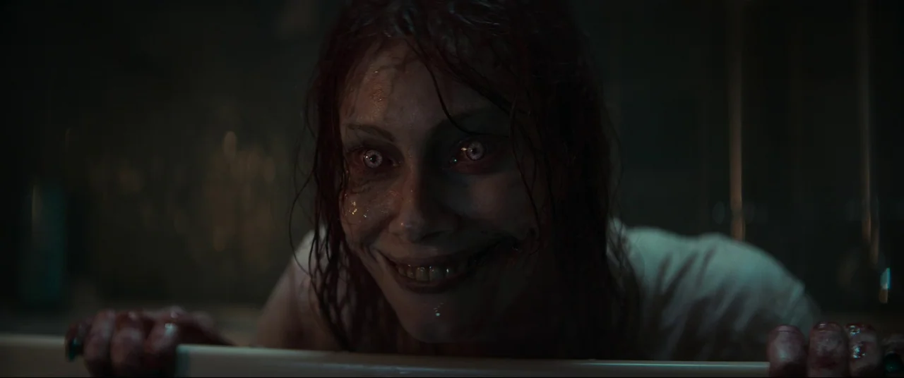 Evil Dead Rise (2023) movie still: closeup — A dimly lit room with a stark white porcelain tub edge in the foreground. A woman with wet hair and…