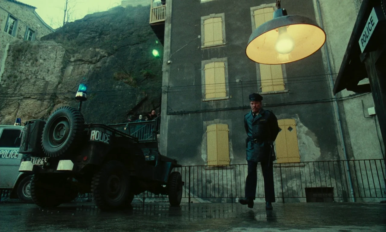Litan (1982) movie still: wide — A stone courtyard sits before a multi-story building with boarded yellow windows at night. A lone police officer…