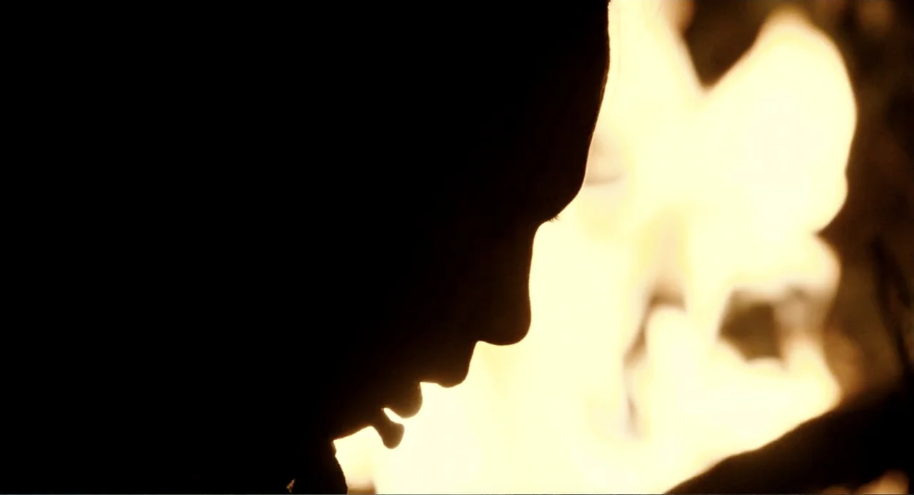 Apocalypto (2006) movie still: closeup — A dark, blurred background is illuminated by the bright, flickering glow of firelight. A woman is seen in…