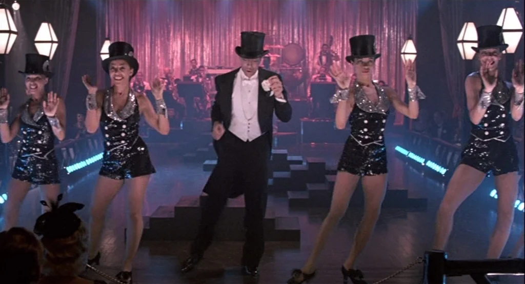 The Cotton Club (1984) movie still: fullbody — A nightclub stage with a metallic red curtain backdrop and tiered wooden levels. A man in formal…