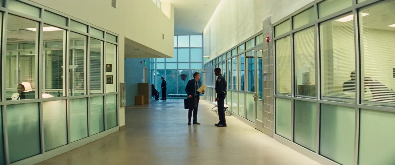 Kinds of Kindness (2024) movie still: wide — A modern police station lobby features glass-walled offices and high ceilings with natural daylight. A…