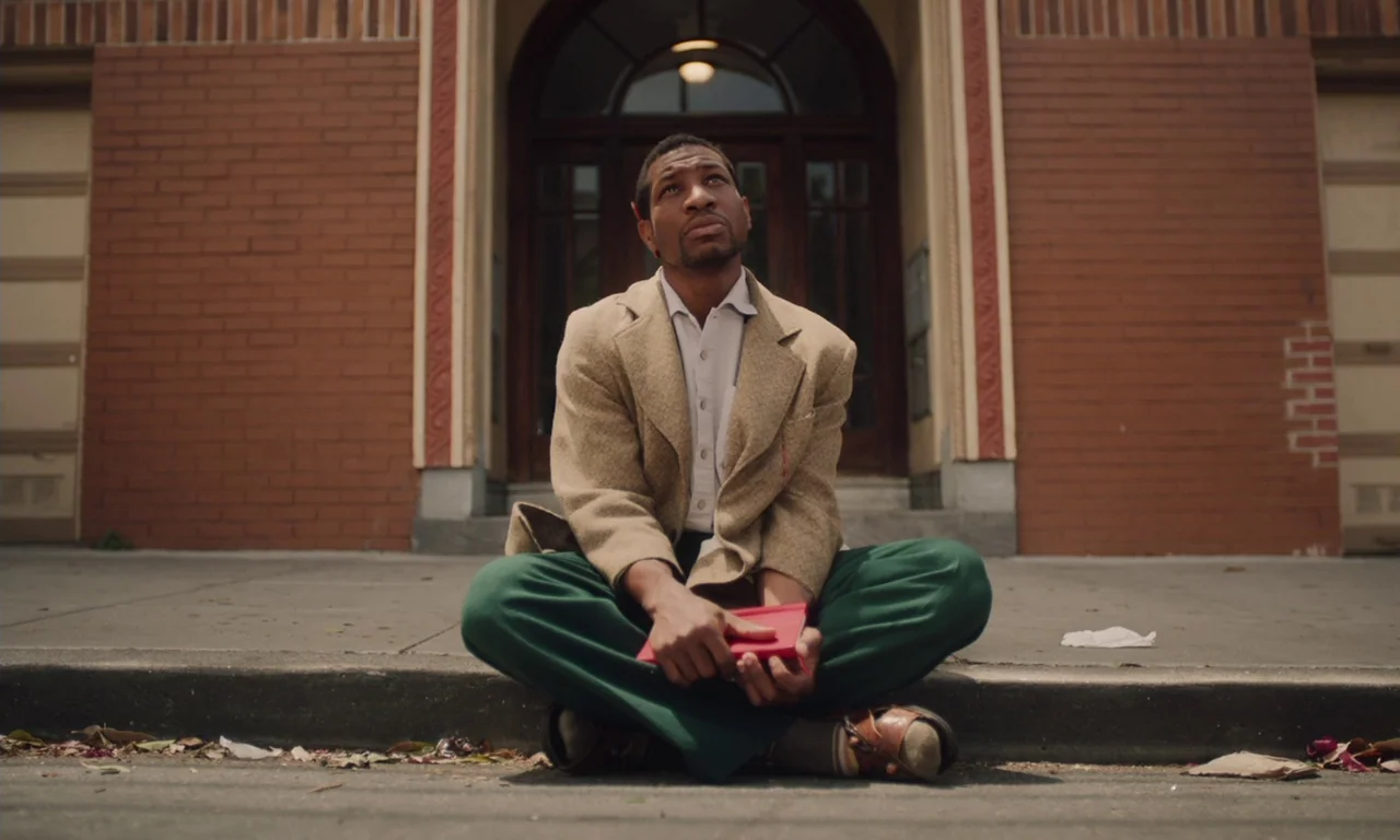 The Last Black Man in San Francisco (2019) movie still: fullbody — A man sits on a concrete sidewalk in front of a brick building entrance. He rests…