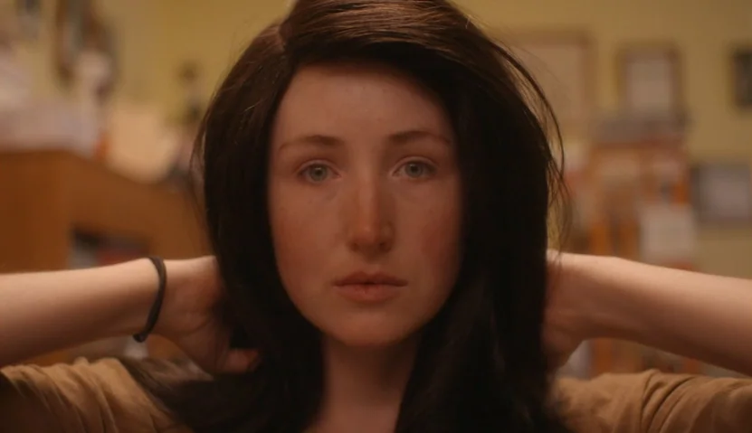 Kate Plays Christine (2016) movie still: closeup — A woman stands in a soft-lit room with blurred shelves in the background. She holds her dark hair…