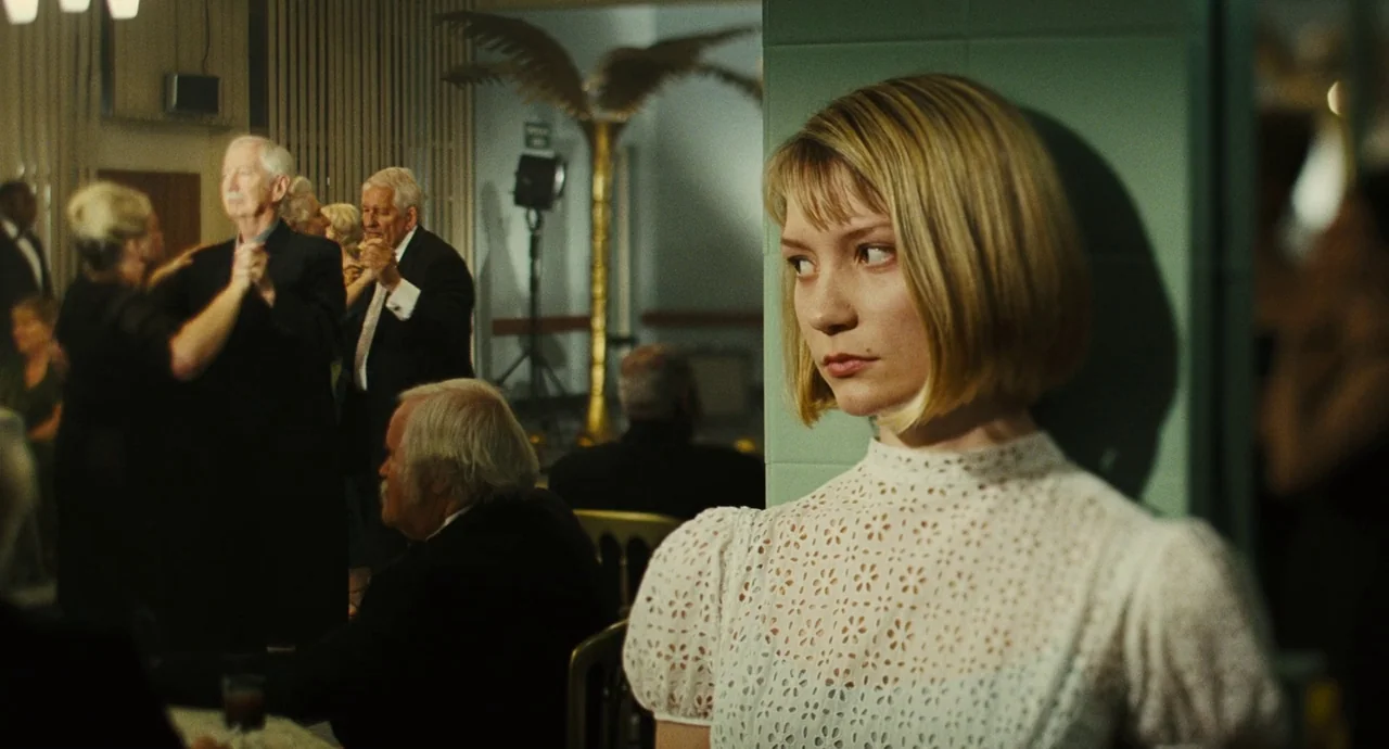 The Double (2013) movie still: medium — A dimly lit ballroom with elderly guests dancing in the background. A woman in a white lace dress stands…