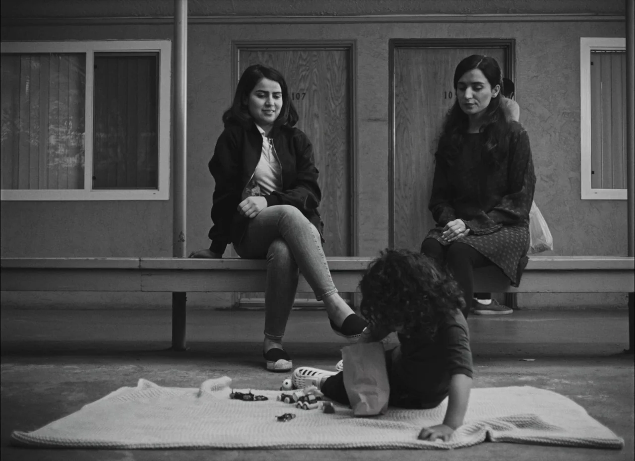 Fremont (2023) movie still: fullbody — A motel exterior features two doors labeled 107 and 108 with a long bench. Two women sit on the bench while a…