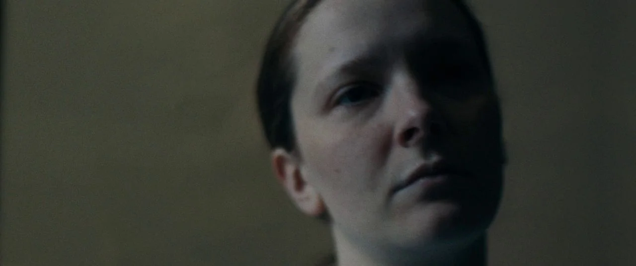 Saint Maud (2019) movie still: closeup — A close-up view of a woman head against a plain, dark background. The subject maintains a steady gaze while…